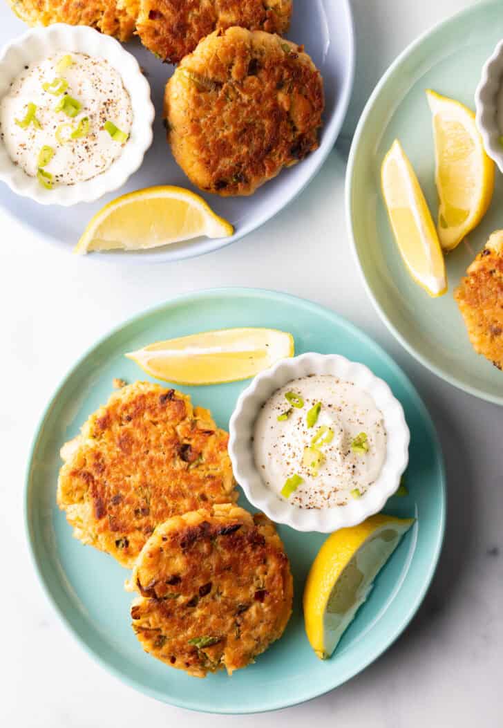 Two salmon patties on a green plate, with a white ramekin of tartar sauce and two lemon wedges.