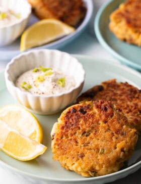 Two salmon patties on a green plate, with a white ramekin of tartar sauce and two lemon wedges.