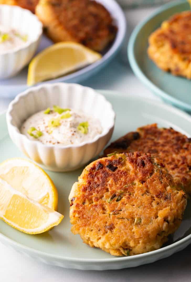 Two salmon patties on a green plate, with a white ramekin of tartar sauce and two lemon wedges.