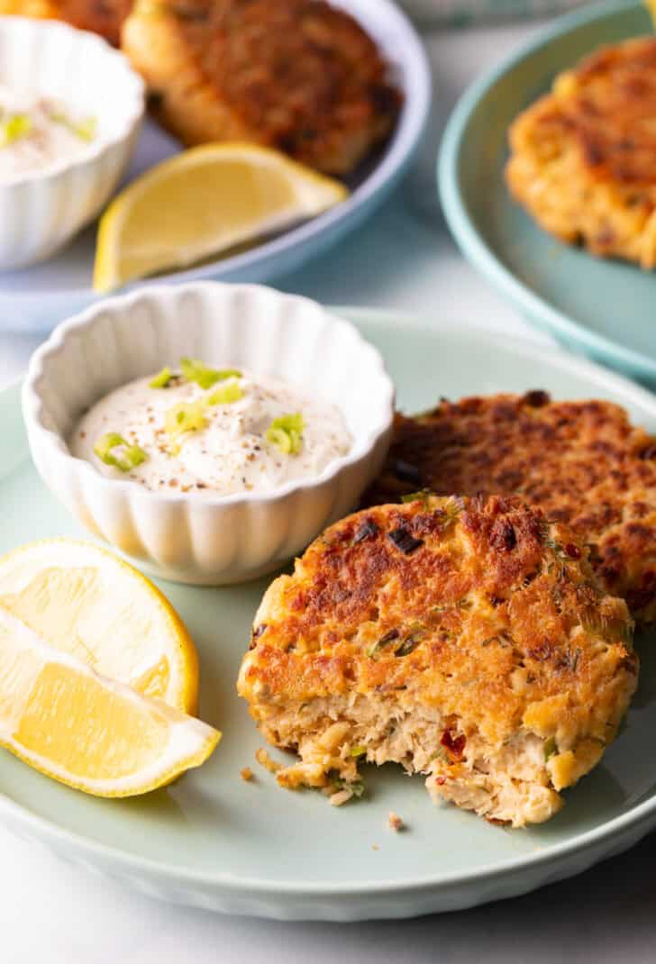 Two salmon patties on a green plate, with a white ramekin of tartar sauce and two lemon wedges. A bite has been taken out of the crab cake closest to camera.