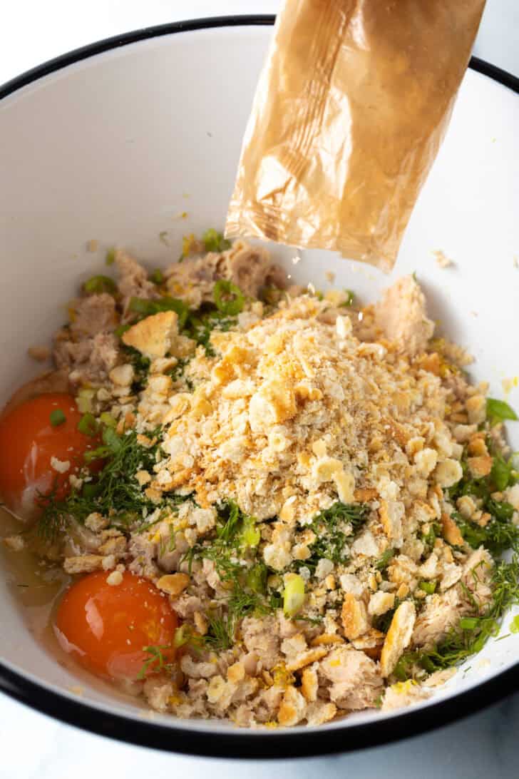 White mixing bowl with canned salmon flakes, breadcrumbs, fresh chopped herbs, and eggs.