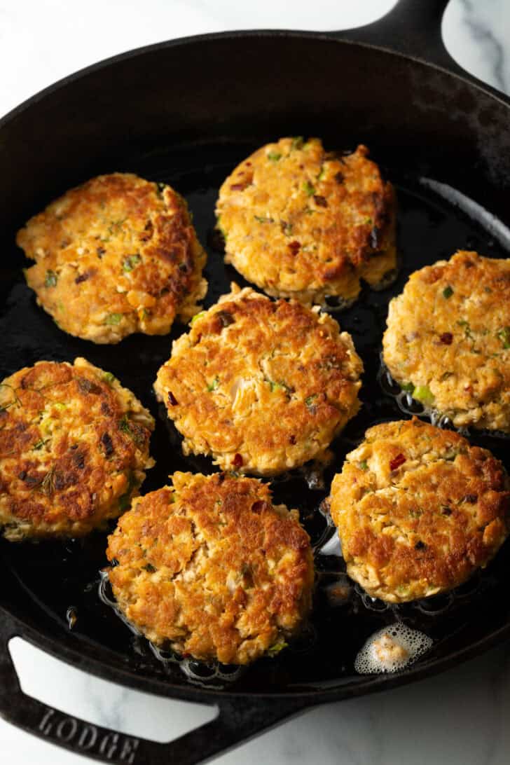 Top down view 7 salmon patties cooking in a cast iron skillet.