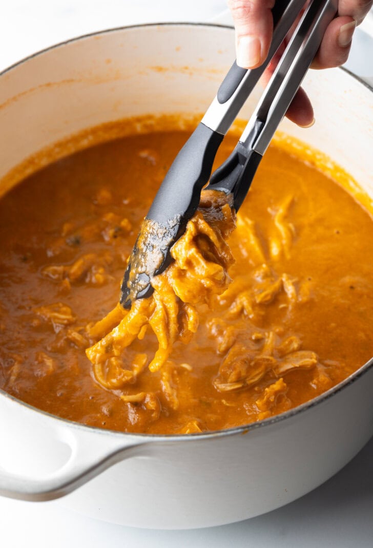 Tongs grabbing at stewed chicken meat in a large pot.