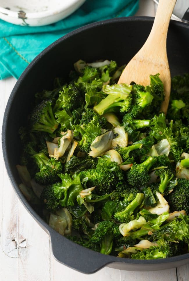 Broccoli, cabbage and kale in a skillet with a wooden spoon stirring it.