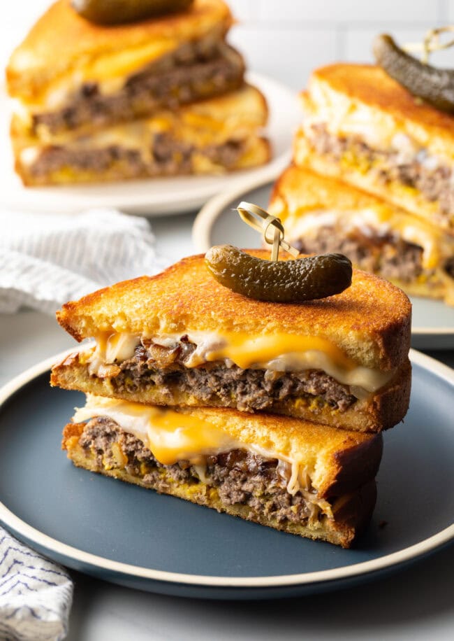 Two halves of a patty melt stacked on top of each other. A wood skewer with small pickle is going through the top of the sandwich stack.