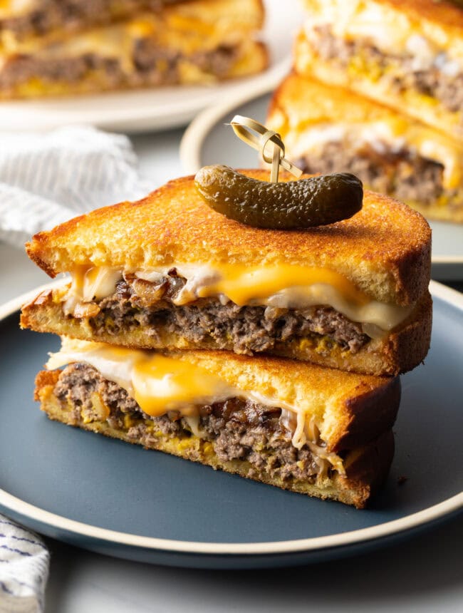 Two halves of a patty melt stacked on top of each other. A wood skewer with small pickle is going through the top of the sandwich stack.