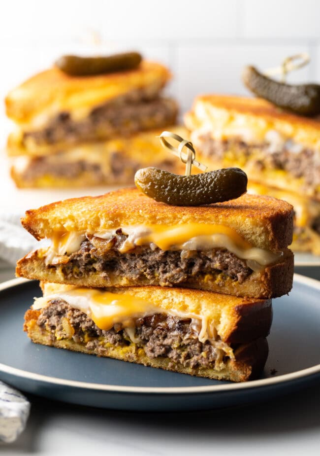 Two halves of a patty melt stacked on top of each other. A wood skewer with small pickle is going through the top of the sandwich stack.