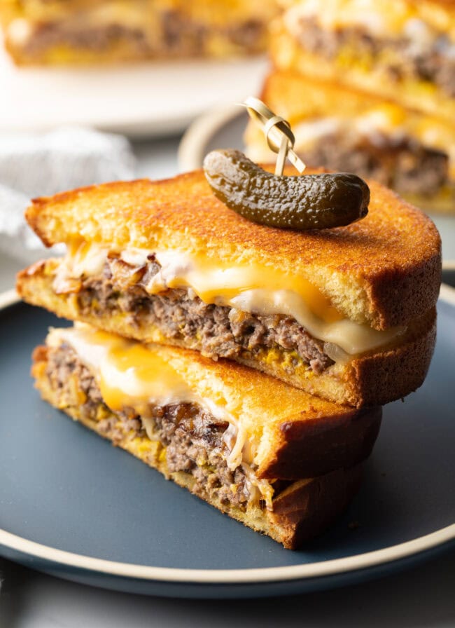 Close view of two halves of a patty melt stacked on top of each other. A wood skewer with small pickle is going through the top of the sandwich stack.