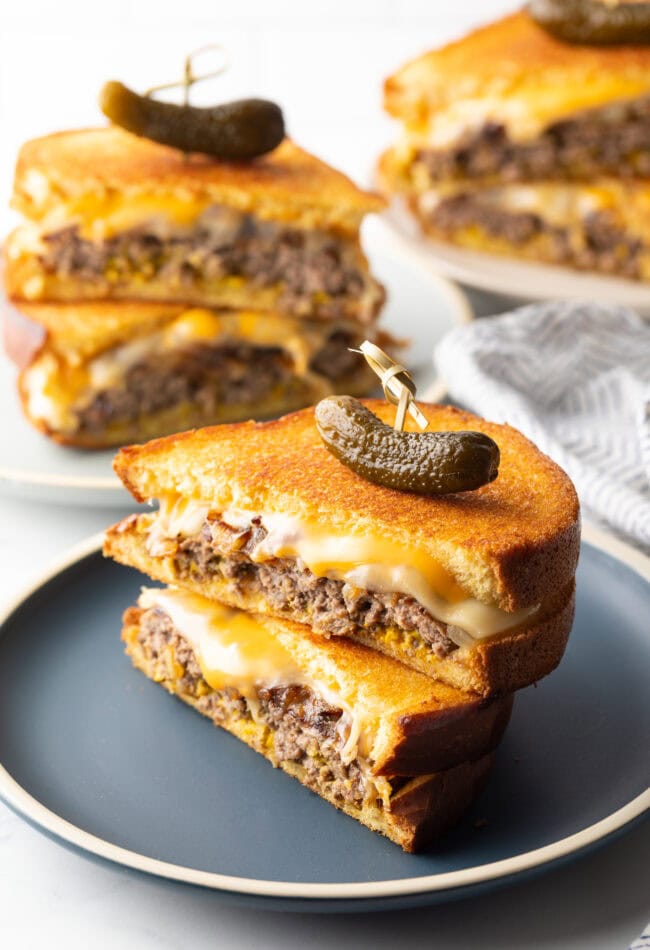 Two halves of a patty melt stacked on top of each other. A wood skewer with small pickle is going through the top of the sandwich stack.