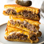 Three pieces of two patty melts stacked on top of each other. A wood skewer with small pickle is going through the top of the sandwich stack.