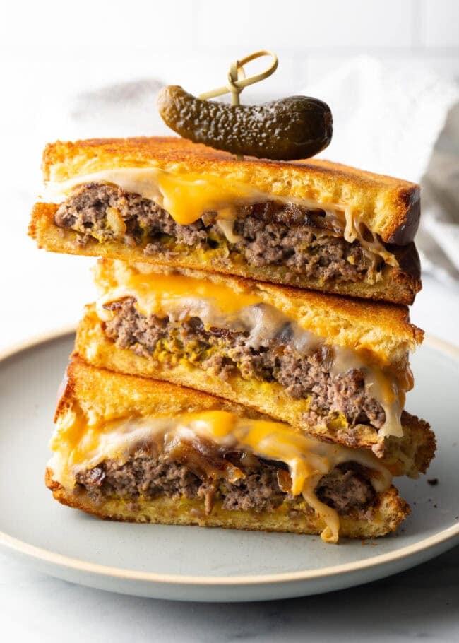 Three pieces of two patty melts stacked on top of each other. A wood skewer with small pickle is going through the top of the sandwich stack.