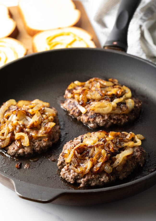 Three burger patties in a skillet, each topped with onion.