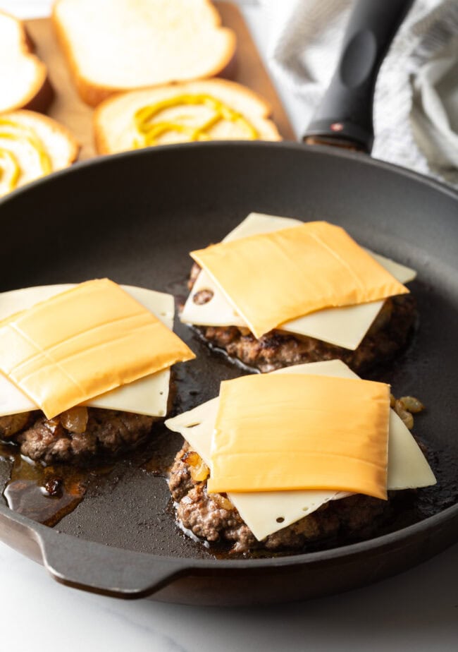 Three burger patties in a skillet with a slice of swiss cheese and slice of cheddar cheese on each.