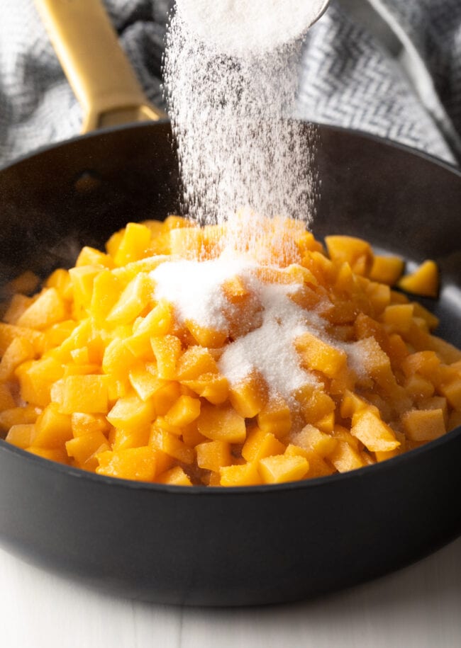 Adding sugar to fresh chopped peaches in a large black pot.