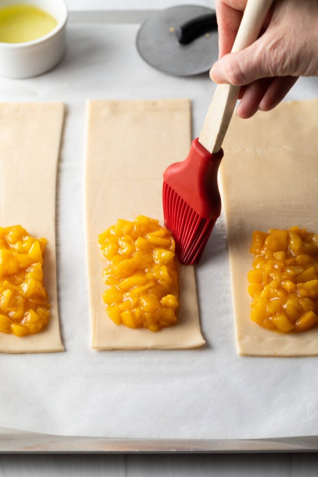 Hand with pastry brush going around outside of the peach filling, getting ready to seal the pie closed.