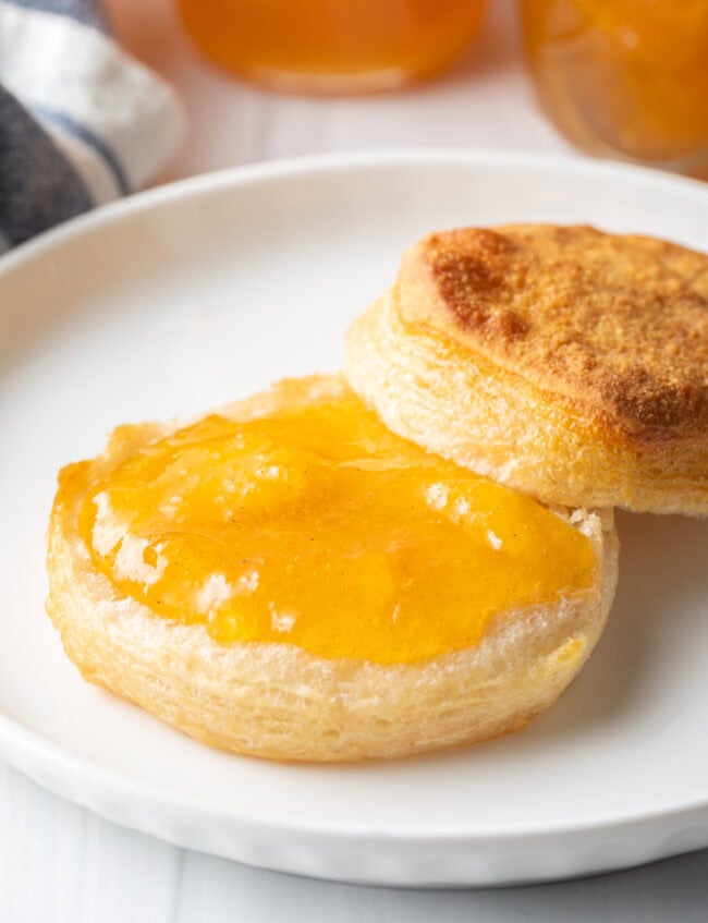 Biscuit split in half on a white plate. Bottom half of biscuit is smeared with peach jam.