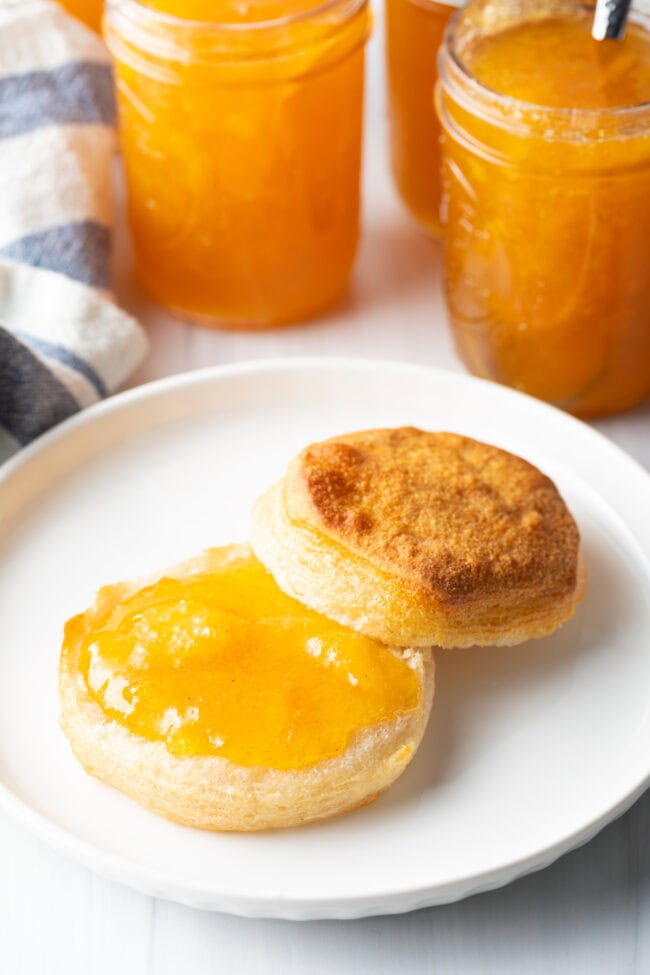 Biscuit split in half on a white plate. Bottom half of biscuit is smeared with peach jam.