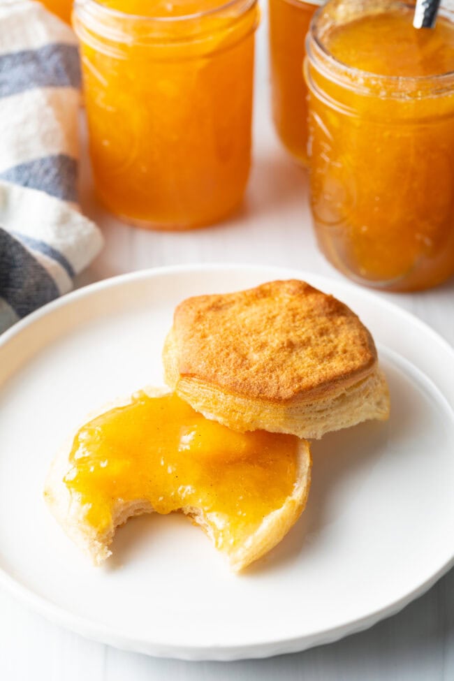 Biscuit split in half on a white plate. Bottom half of biscuit is smeared with peach jam, and has a bite taken out.