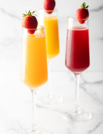 Two champagne flutes filled with orange mimosa, and another glass filled with a cranberry mimosa. Both glasses are garnished with a whole strawberry on the rim.