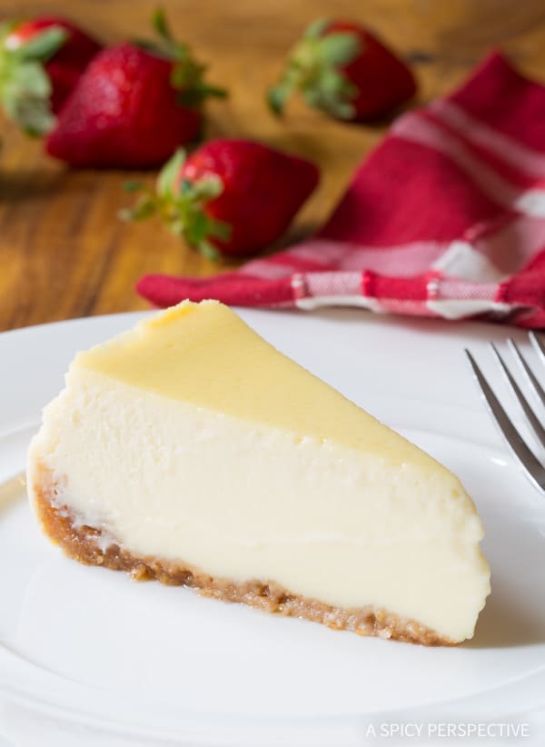 A slice of slow cooker cheesecake on a white plate with strawberries in the background.