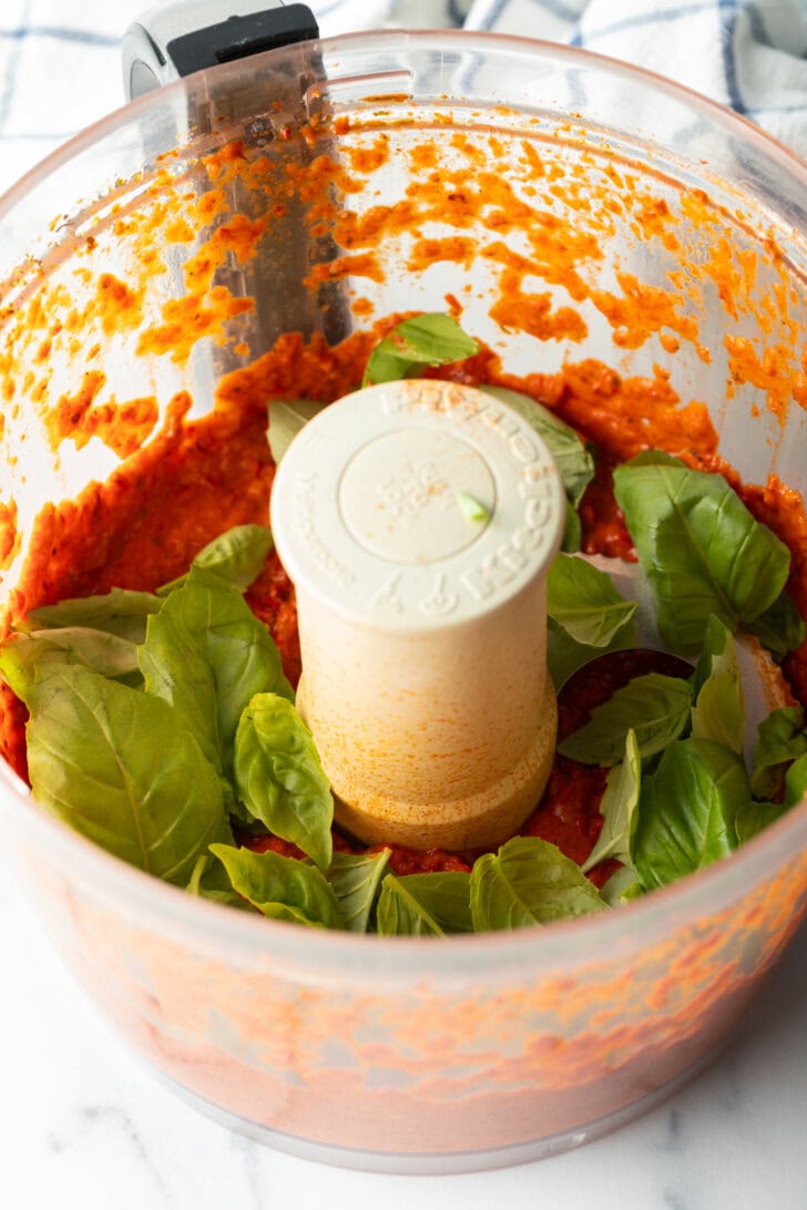 Adding fresh basil to the pureed red pepper sauce in a food processor.