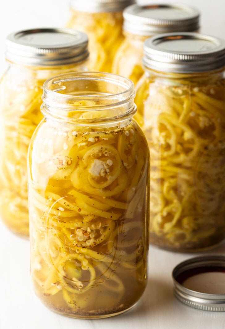 4 large jars of canned pickled banana peppers. The jar closest to the camera has the lid off showing the slices of peppers filling the jar.