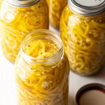 4 large jars of canned pickled banana peppers. The jar closest to the camera has the lid off showing the slices of peppers filling the jar.