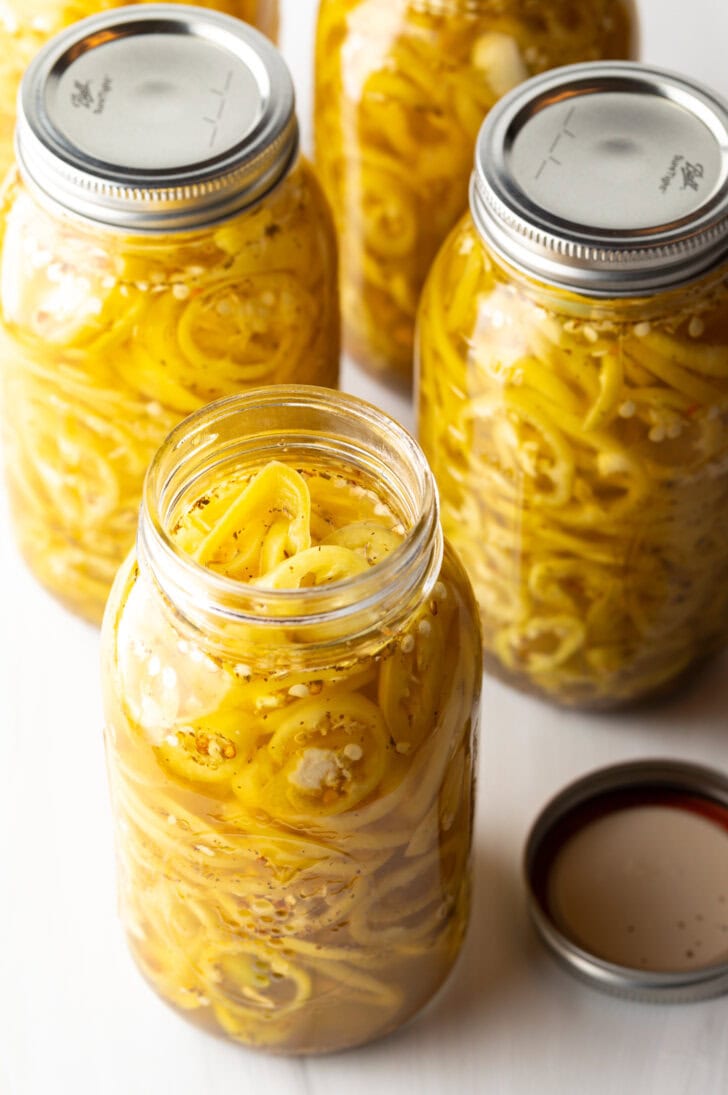 4 large jars of canned pickled banana peppers. The jar closest to the camera has the lid off showing the slices of peppers filling the jar.