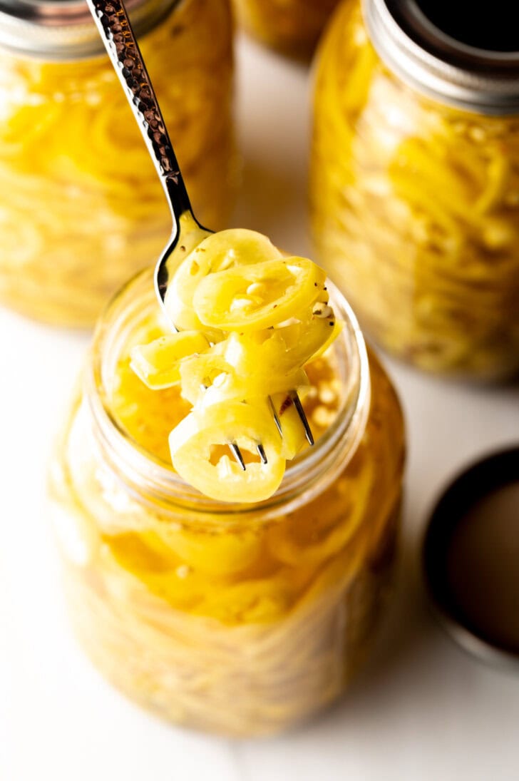 4 large jars of canned pickled banana peppers. The jar closest to the camera has the lid off showing the slices of peppers filling the jar. A fork is pulling a serving of slices from the jar.