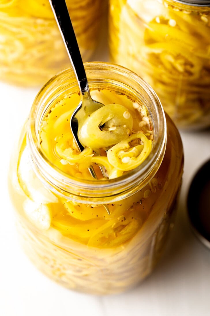 4 large jars of canned pickled banana peppers. The jar closest to the camera has the lid off showing the slices of peppers filling the jar. A fork is pulling a serving of slices from the jar.