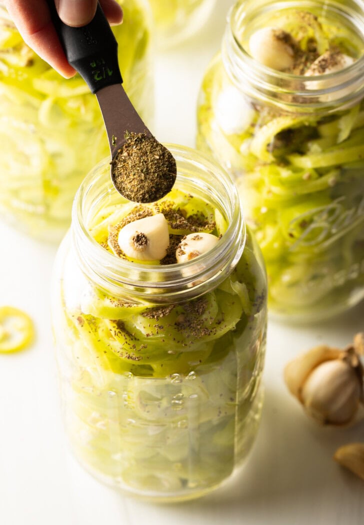 Adding spices to glass jars filled with pepper slices and garlic cloves.