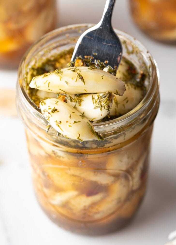 Top down open glass jar stuffed with preserved garlic and herbs. A fork is digging in to grab several cloves.