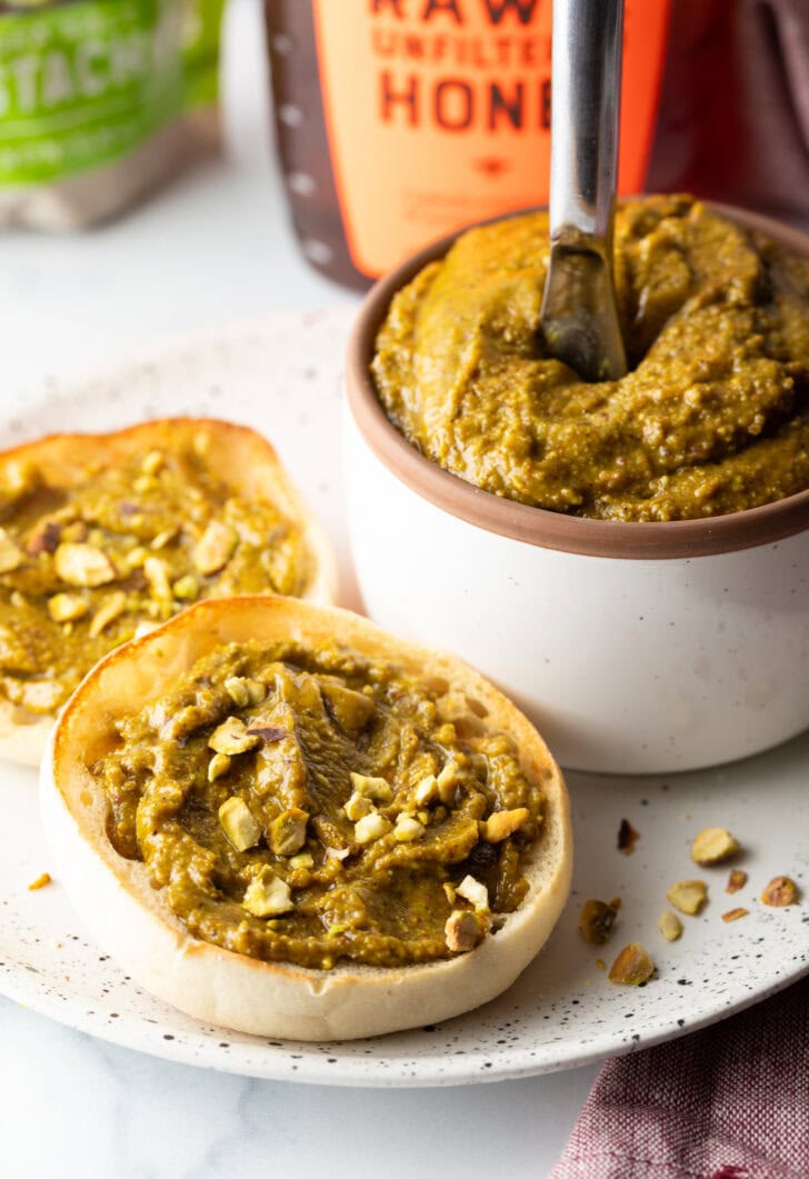 White bowl with pistachio butter. To the left side are two english muffin halves, each smeared with the butter and topped with fresh chopped nuts.