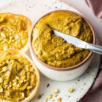 Top down view of a white bowl with pistachio butter, with a metal knife running through it. To the left side are two english muffin halves, each smeared with the butter and topped with fresh chopped nuts.