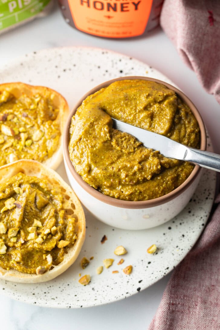 Top down view of a white bowl with pistachio butter, with a metal knife running through it. To the left side are two english muffin halves, each smeared with the butter and topped with fresh chopped nuts.