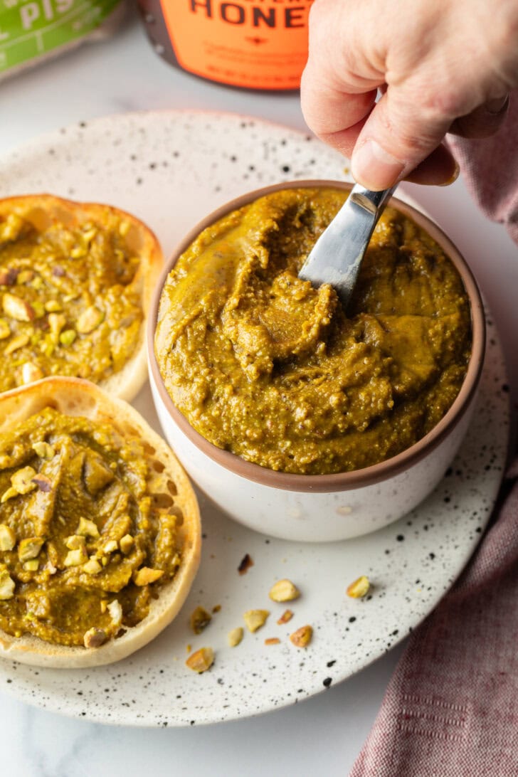 Top down view of a white bowl with pistachio butter, a hand with a metal knife running through it. To the left side are two english muffin halves, each smeared with the butter and topped with fresh chopped nuts.