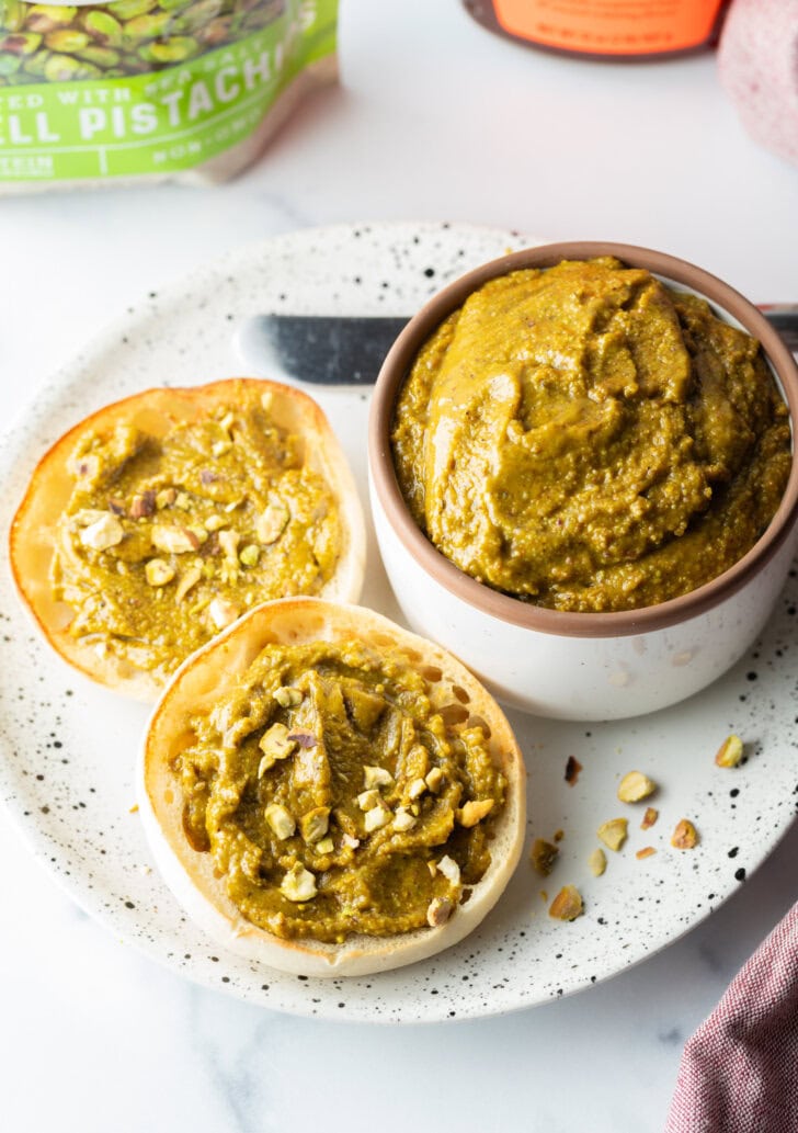 Top down view of a white bowl with pistachio butter. To the left side are two english muffin halves, each smeared with the butter and topped with fresh chopped nuts.