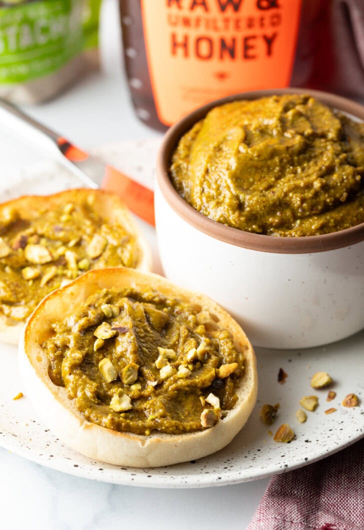 white bowl with pistachio butter. To the left side are two english muffin halves, each smeared with the butter and topped with fresh chopped nuts.