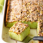 A slice of pistachio pudding poke cake on a spatula resting in the rectangle pan, showing to camera.