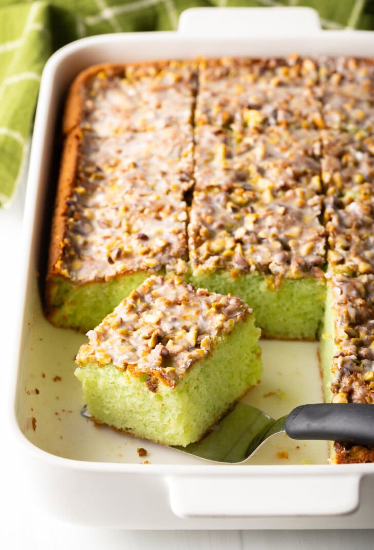 A slice of pistachio pudding cake on a spatula resting in the rectangle pan, showing to camera.