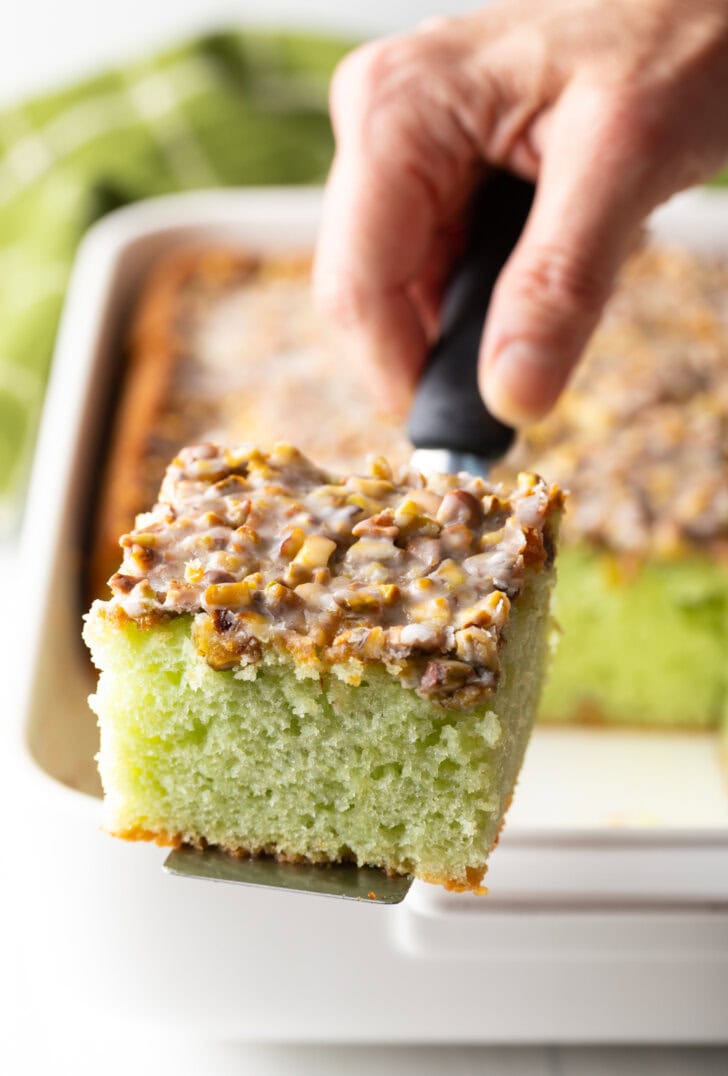 Hand with a slice of pistachio pudding poke cake on a spatula, showing to camera.