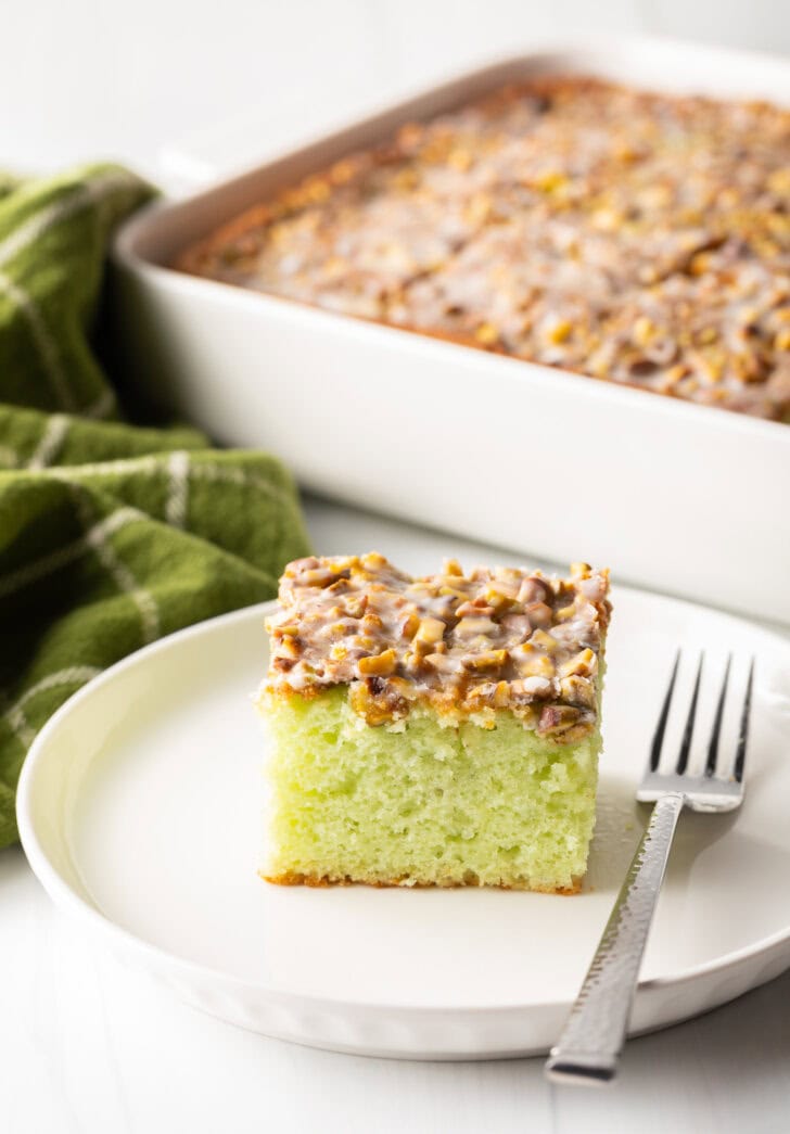 Square piece of pistachio pudding cake on a white plate, with fork next to the slice.