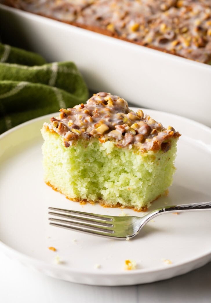 Square piece of pistachio pudding cake on a white plate, with fork next to the slice. A bite has been taken from the cake.