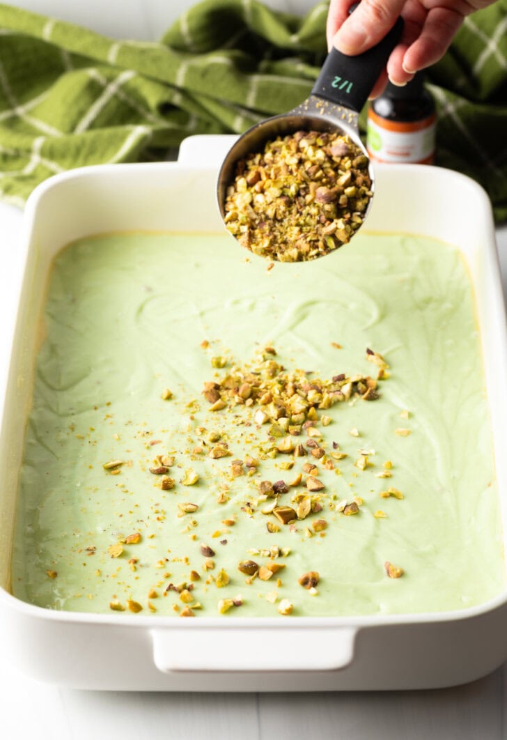 Measuring scoop sprinkling chopped pistachios over the green cake batgle pan.ter in a rectan