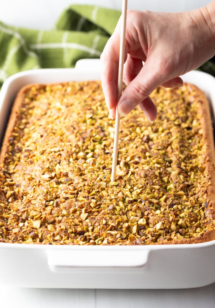 Baked pudding cake with pistachios. A hand with a wooden skewer is poking holes in the top of the cake.