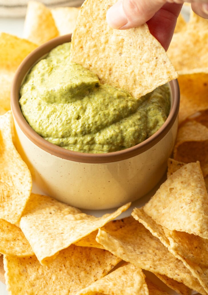 Hand scooping a tortilla chip into a bowl of poblano cream sauce.