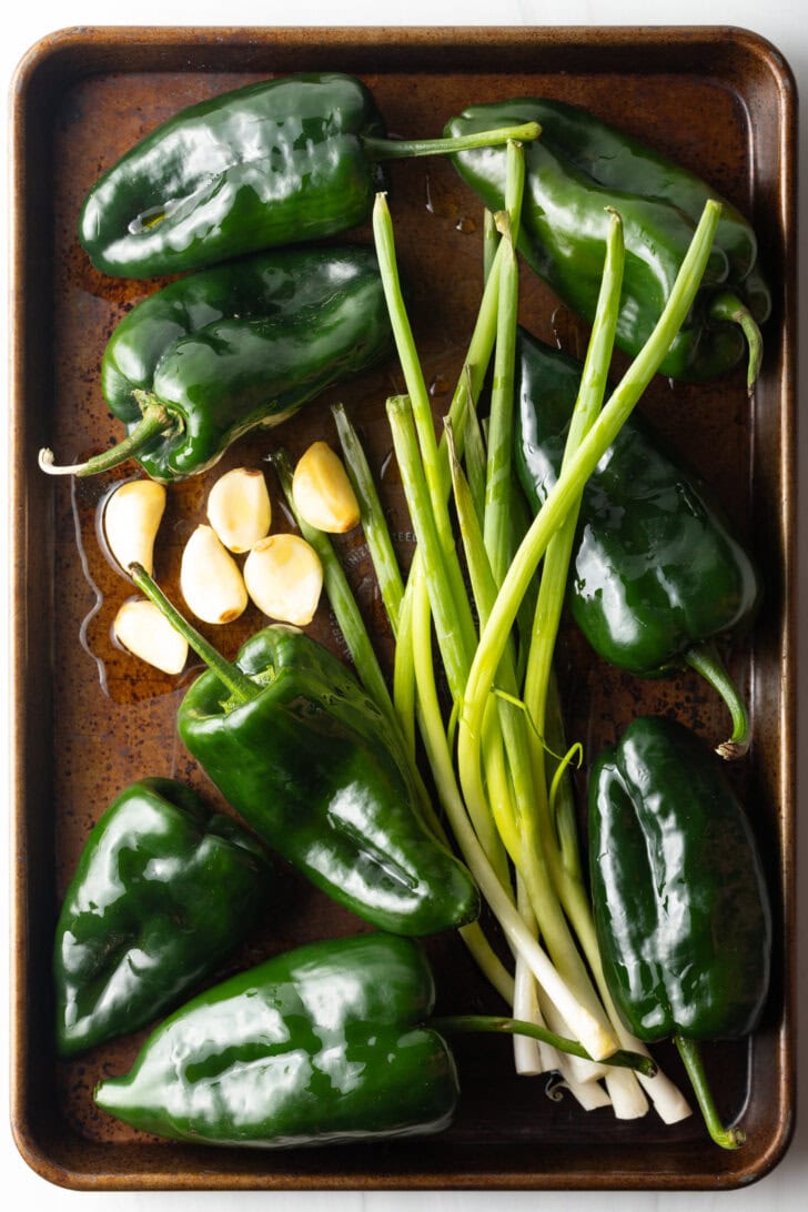 Recipe ingredients: 8 raw whole poblano peppers on a roasting pan with whole green onions, cloves of peeled garlic.