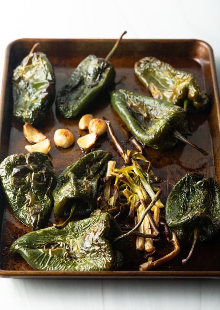 8 whole roasted poblano peppers on a roasting pan with whole green onions, cloves of peeled garlic.