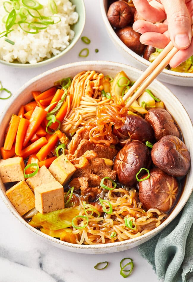 Overhead shot of bowl of sukiyaki with pork recipe with chopsticks on top pulling noodle up