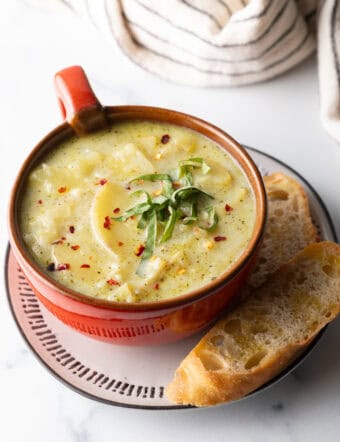 Red oversized mug filled with leek and potato soup, on a plate with slice of baguette on the side.